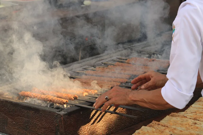 Eine Person wendet einen Adana-Spieß mit perfekt gegrilltem Hackfleisch auf einem Holzkohlegrill.