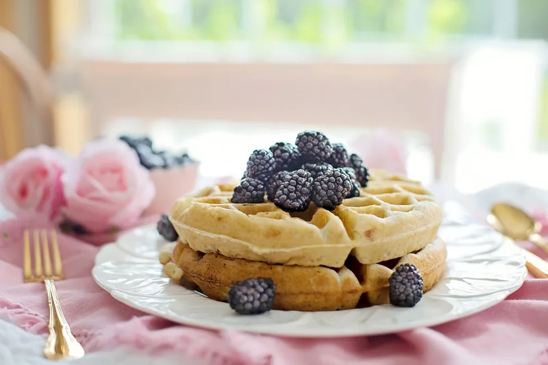 Eine Hand gießt frischen Waffelteig in ein heißes belgisches Waffeleisen. Zeigt den Backvorgang in der Küche.
