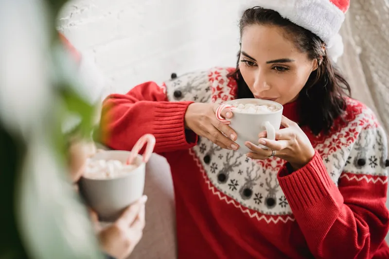 Gemütliche Weihnachtsszene im Wohnzimmer, mit einer Frau im Hintergrund, die einen lustigen Weihnachtspullover trägt.
