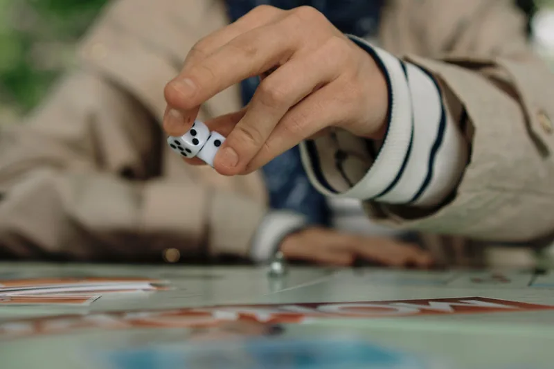 Eine Person würfelt gerade auf einem Brettspiel, wobei der Fokus auf der Hand und den Würfeln liegt.