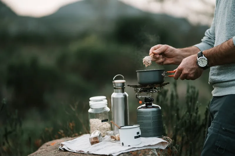 Eine Person rührt in einem Topf auf einem Campingkocher und bereitet ein Gericht in der Natur zu.