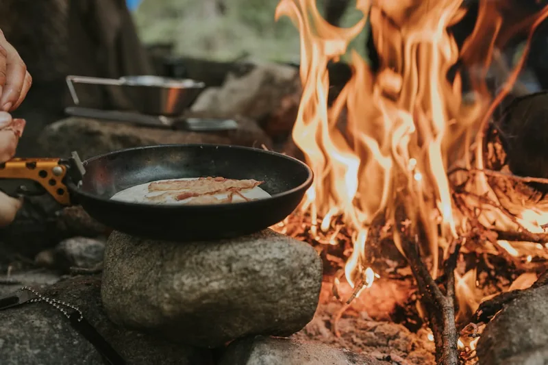 Gemütliche Campingszene mit frisch zubereitetem Essen auf dem Tisch neben einem Lagerfeuer.