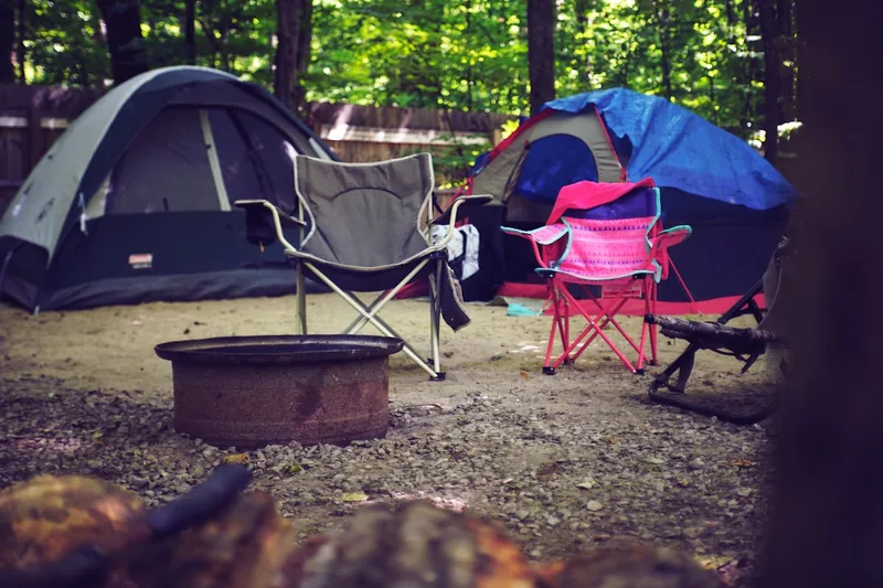 Idyllische Campingszene bei Sonnenuntergang mit mehreren Campingstühlen um eine Feuerstelle.