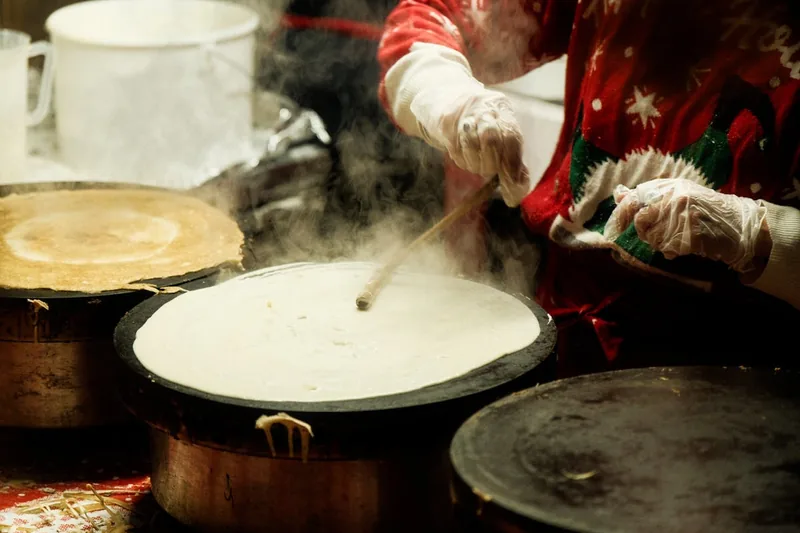 Eine Hand verteilt Crêpe-Teig in einer flachen Crêpes-Pfanne auf dem Herd, in Aktion beim Backen.