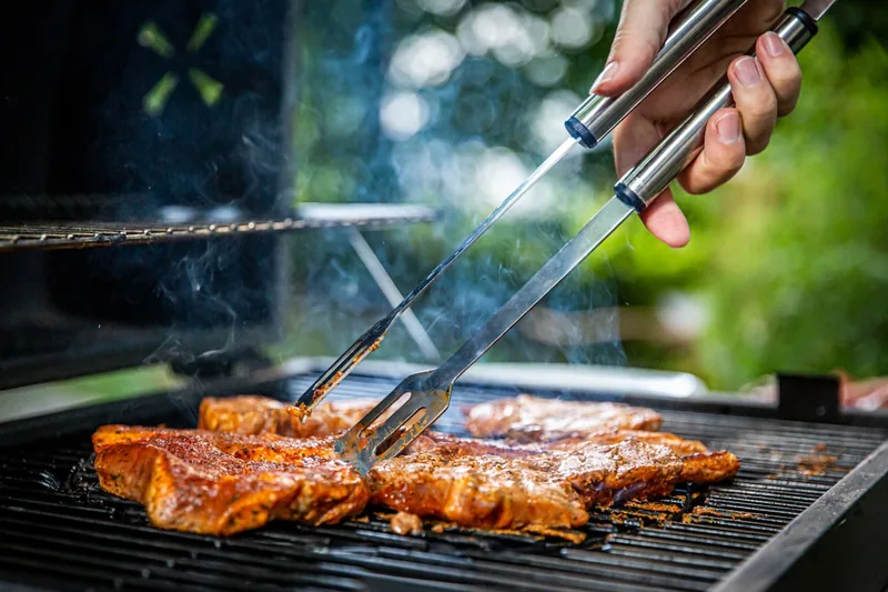 Eine Person wendet mit einer Grillzange ein Steak auf dem heißen Rost eines Gasgrills.