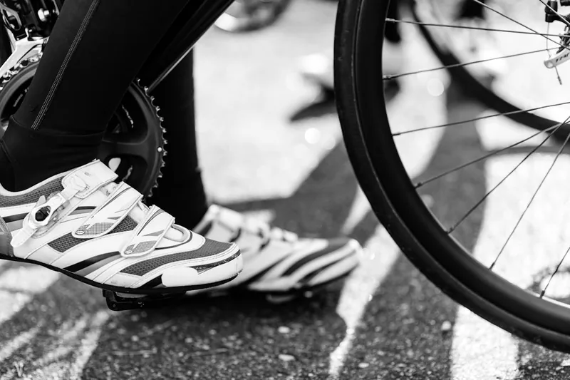 Eine Person tritt in die Fahrradpedale; ein Fuß in Fahrradschuh ruht auf dem Pedal.
