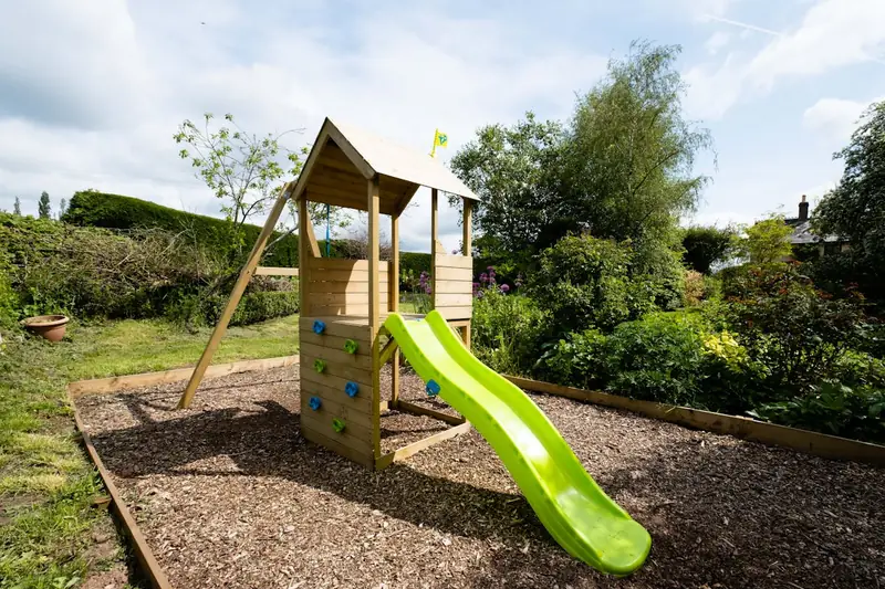 Ein hochwertiger Holz-Spielturm mit Rutsche und Schaukel vor leicht verschwommenem Gartenhintergrund.