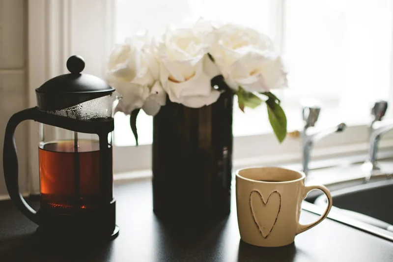 Eine dampfende Tasse Kaffee auf einem Küchentresen am Morgen, French Press im Hintergrund.