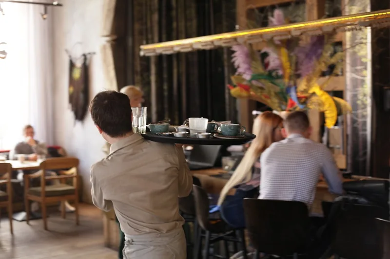 Ein Kellner trägt ein volles Gastro-Tablett mit Speisen und Getränken in einem geschäftigen Restaurant.