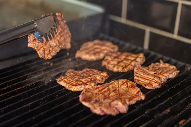 Eine Hand wendet mit einem Grillwender saftiges Fleisch auf einem heißen Holzkohlegrill.