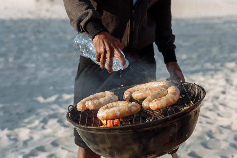 Eine Person grillt Würstchen auf einem kompakten Grilleimer, Rauch steigt auf, Fokus auf Nutzung.