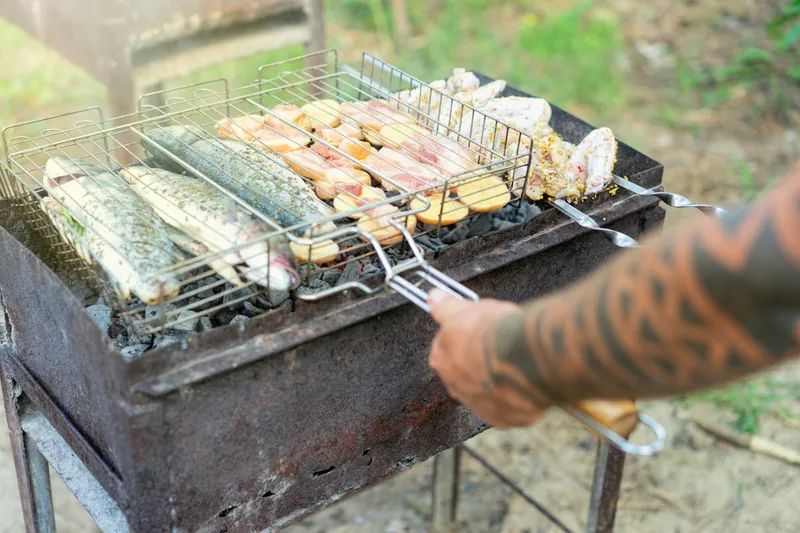 Eine Person wendet saftiges Grillgut auf einem Holzkohlegrill, Funken fliegen, Rauch steigt auf.