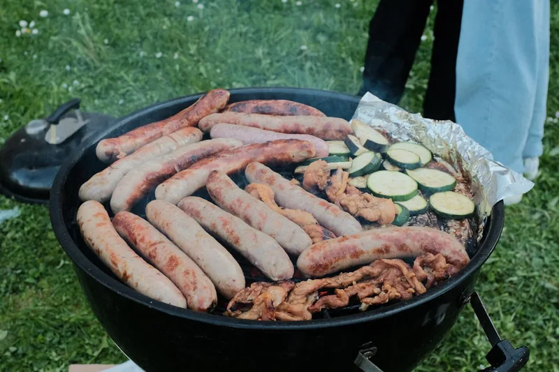 Gemütliche Gartenparty-Szene mit Freunden und Familie im Hintergrund, im Vordergrund ein leicht rauchender Holzkohlegrill, der eine warme Atmosphäre schafft.