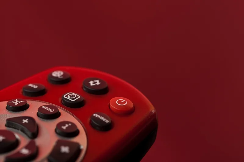 Eine Hand hält eine Fernbedienung und bedient eine IPTV-Box, im Hintergrund ist ein Fernsehbildschirm angedeutet.