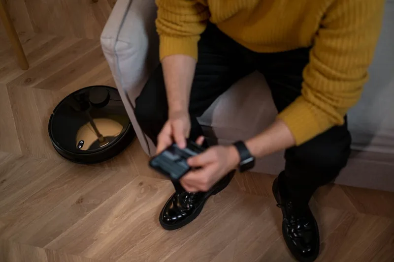 Eine Hand hält ein Smartphone mit einer Saugroboter-App, im Hintergrund fährt der Staubsaugroboter.