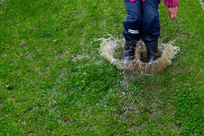 Ein Kind mit farbenfrohen Gummistiefeln planscht fröhlich in einer großen Pfütze, Fokus auf die Stiefel.