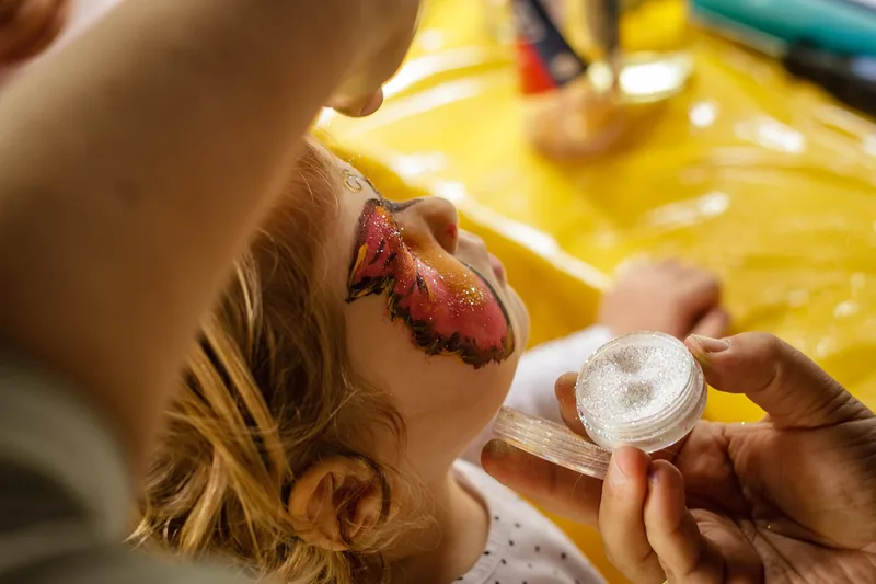 Eine Erwachsene bemalt vorsichtig das Gesicht eines Kindes mit bunter Kinderschminke, Nahaufnahme der Anwendung.