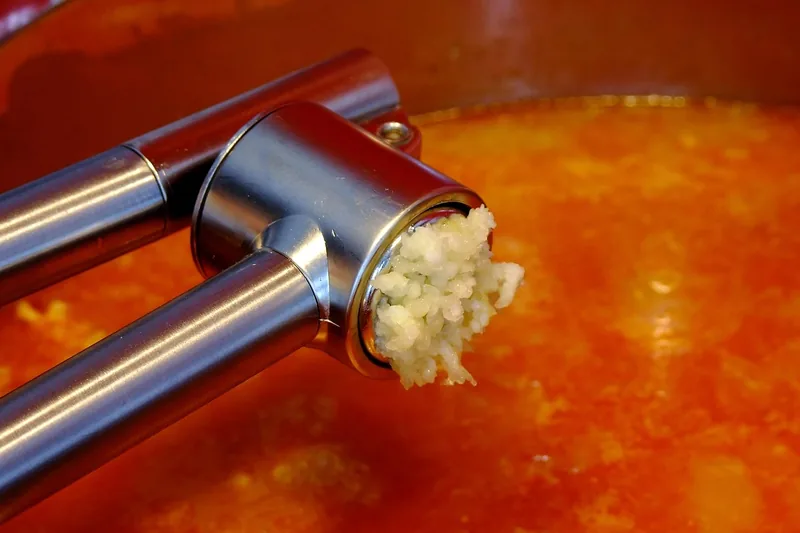 Eine Hand presst Knoblauchzehen mit einer modernen Knoblauchpresse in eine Schale, die Anwendung zeigend.
