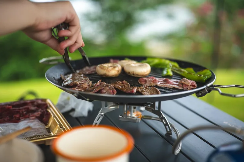 Eine entspannte Outdoor-Grillszene mit einem Koffergrill im Hintergrund, während Freunde das Essen am Seeufer genießen.