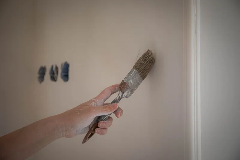 Eine Hand streicht eine Wand mit einem Malerpinsel, zeigt die praktische Anwendung des Werkzeugs beim Renovieren.