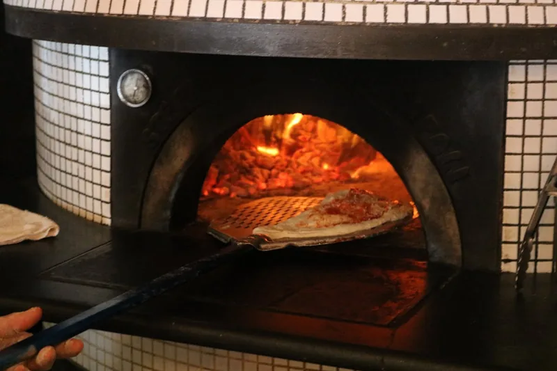 Eine Person schiebt eine frische Pizza mit dem Pizzaschieber in den heiß glühenden Ofen.
