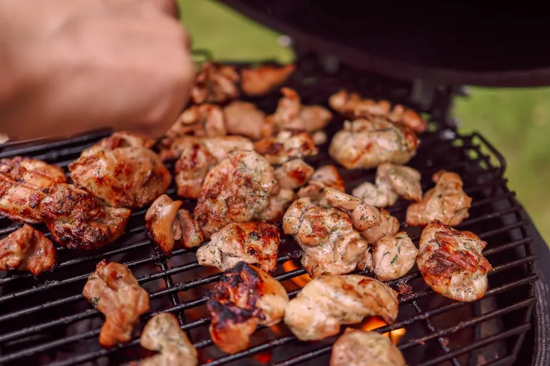 Eine Person legt Grillgut auf den Rost eines glühenden Rundgrills, Rauch steigt auf.