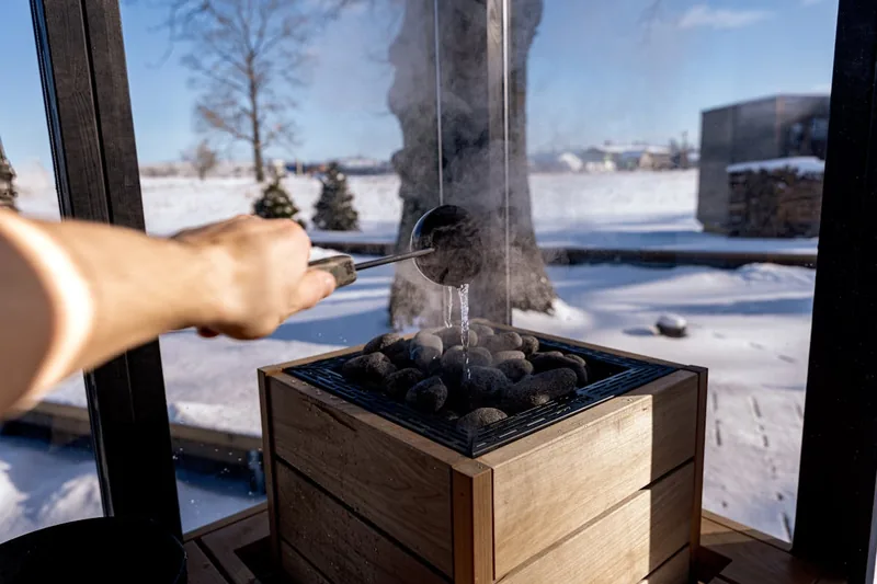 Eine Hand gießt Wasser auf die heißen Saunasteine eines Saunaofens, wodurch effizient Dampf für die Sauna entsteht.