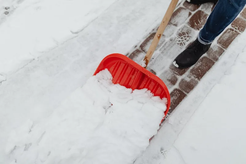 Eine Person schiebt Schnee von einem gepflasterten Gehweg mit einem Schneeschieber, aktiv in Nutzung.
