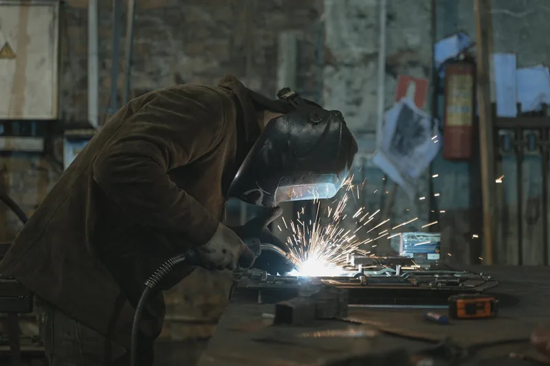 Eine Person schweißt Metall in einer Werkstatt, mit hellen Funken und Schweißhelm, fokussiert auf die Arbeit.