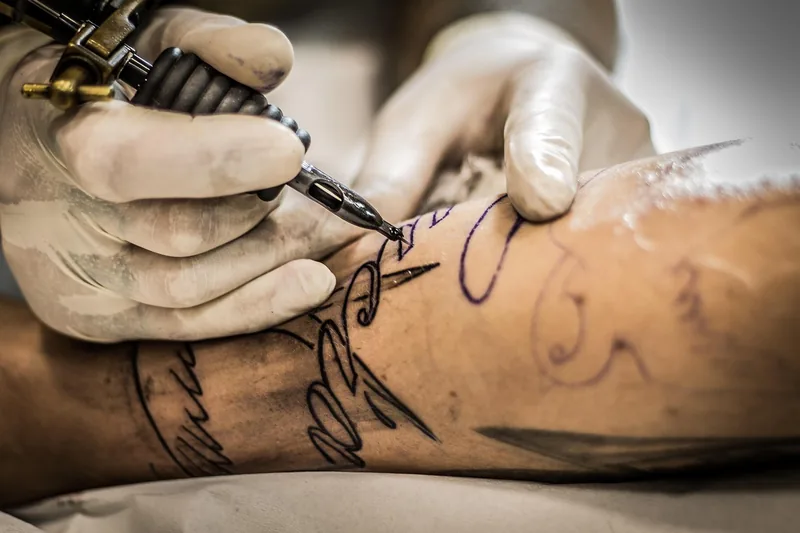 Eine Person taetowiert mit einem Tattoo-Pen einen Arm, Fokus auf die Anwendung und die geuebte Hand.