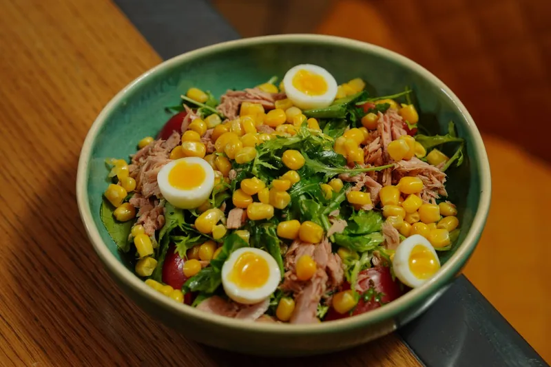 Eine Hand gibt Thunfisch aus der Dose in eine Salatschuessel, frische Zutaten sichtbar.