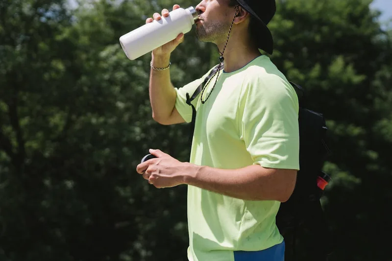 Eine Person trinkt während einer sommerlichen Wanderung aus einer Sporttrinkflasche im Wald.