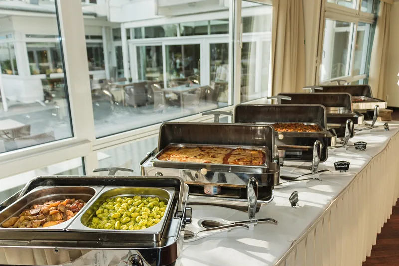 Eine Hand platziert eine elegant angerichtete Schale mit warmen Speisen auf einer Warmhalteplatte an einem Buffet.