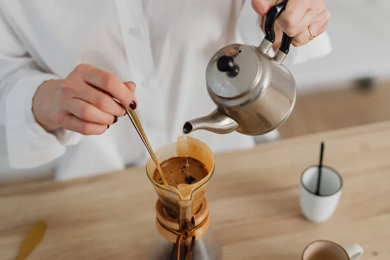 Eine Person gießt heißes Wasser aus einem Edelstahl-Wasserkocher in eine Tasse.